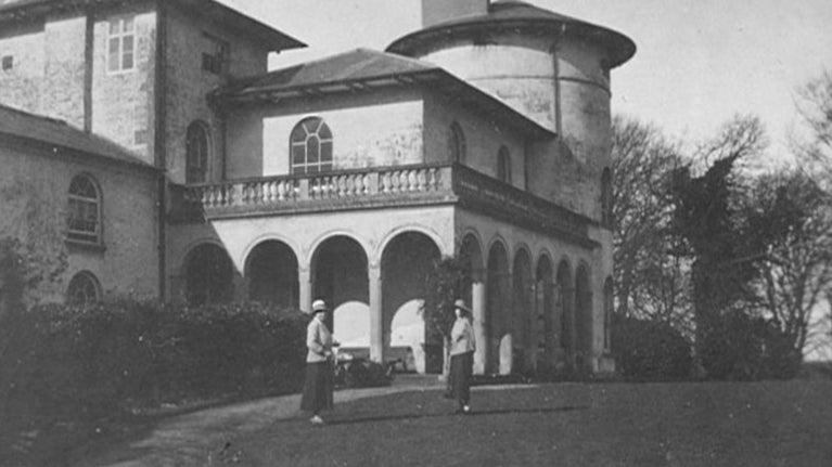 This is a historic black and white photograph showing two women standing in front of a large, elegant house. The building features a rounded tower on the right, arched windows, and a balcony with balustrades. Below the balcony are a series of arches, forming an open gallery or veranda. The women are dressed in early 20th-century attire, wearing long skirts and hats. They are standing on a grassy lawn, with trees visible in the background. The house is surrounded by greenery, and the overall scene conveys a peaceful, historical setting.